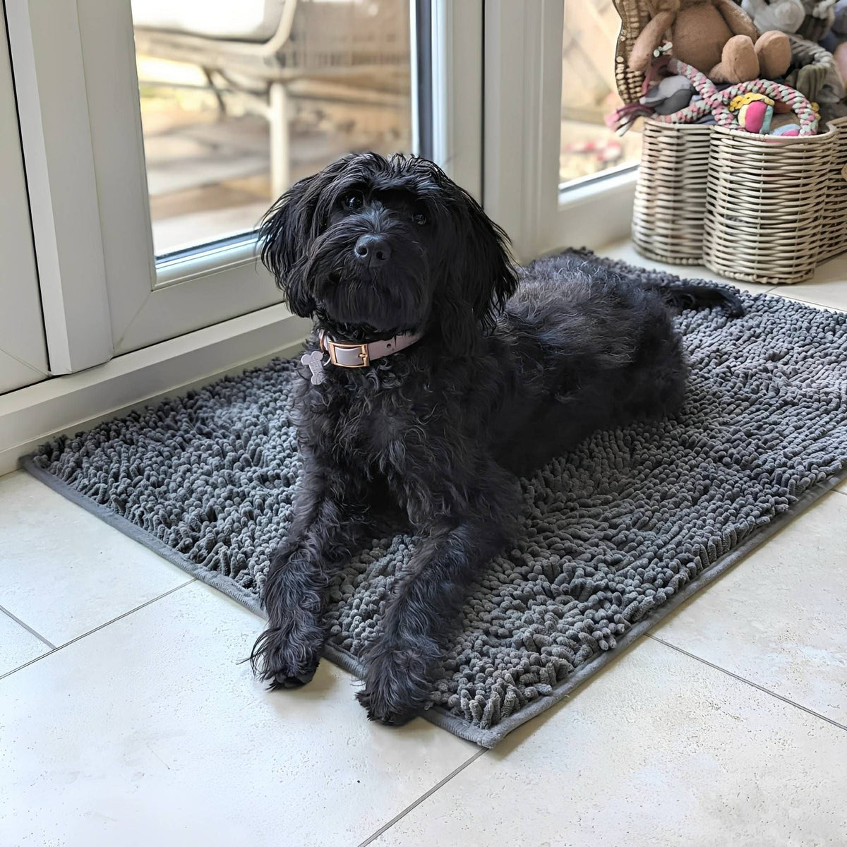 Black shaggy dog in a pink collar lying on a dark grey chenille mat on tiles beside a glazed door. 3