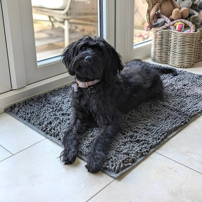 Black shaggy dog in a pink collar lying on a dark grey chenille mat on tiles beside a glazed door. 3