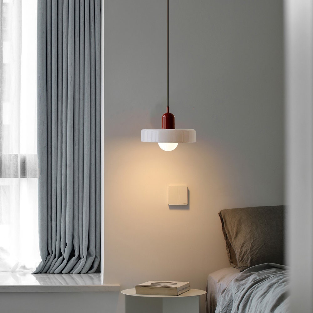 Lit white glass pendant with a burgundy cap casting warm light beside a bed and grey curtains 37
