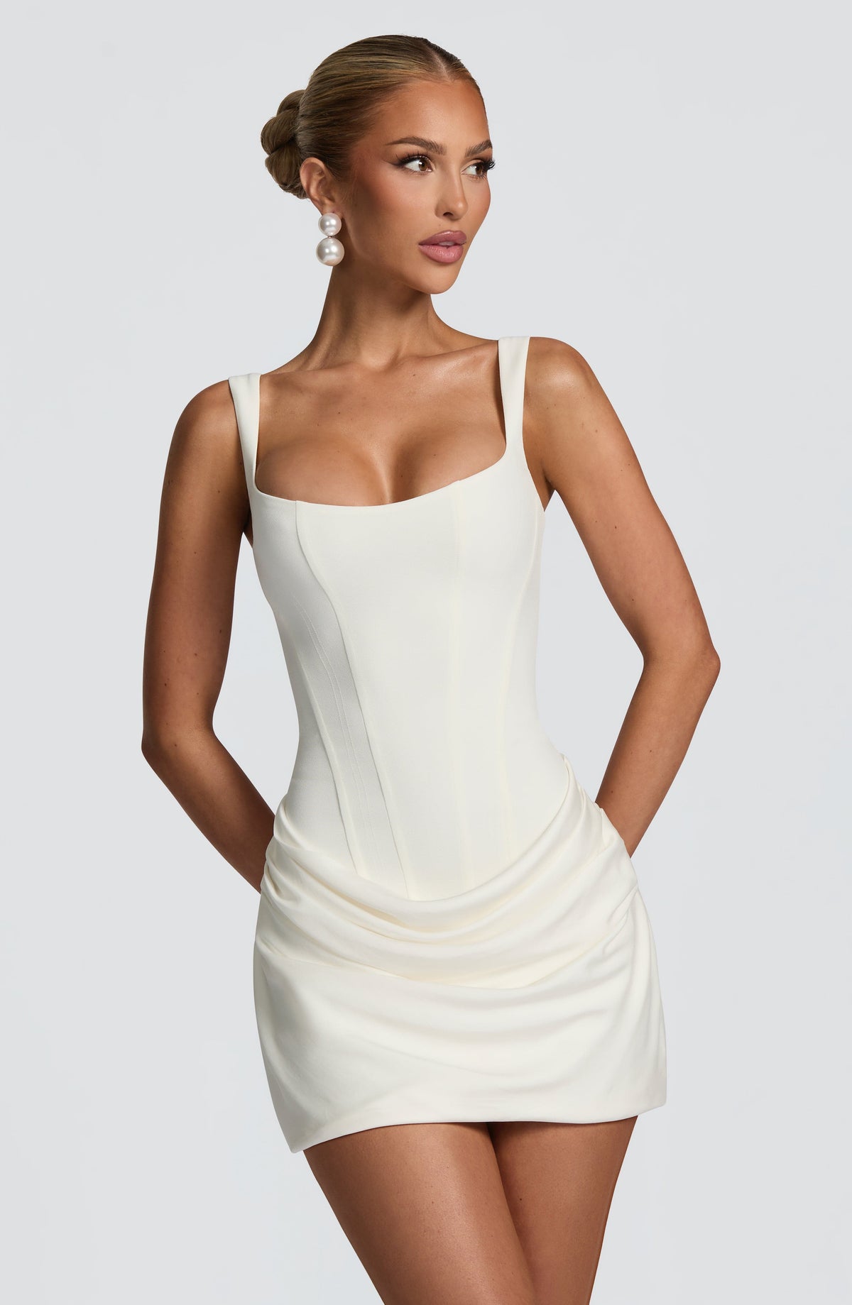 Close front view of the cream mini dress with square neckline, corset seams and draped skirt, worn with pearl earrings. 5