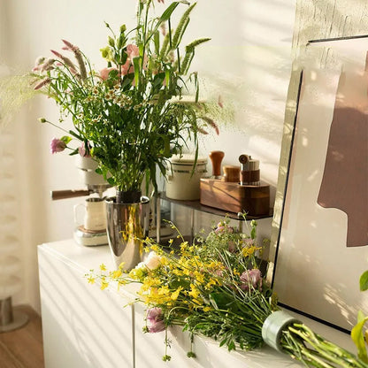 Wild flower arrangement in a metal vase on a white sideboard, with another bouquet lying beside it, its stems bound by a pale green insert. 3