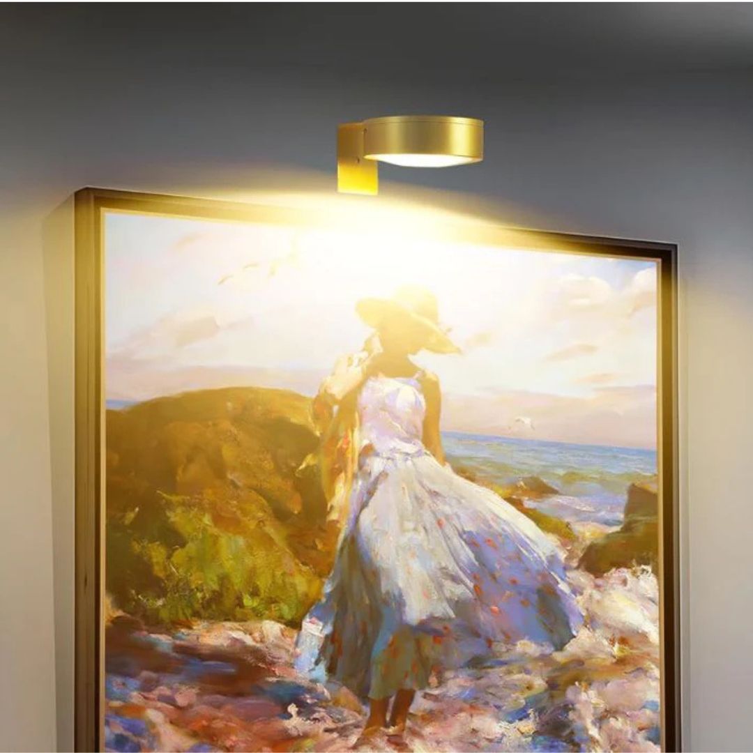 Brass-coloured round light mounted above a framed seaside painting of a woman in a sun hat, casting warm light 1