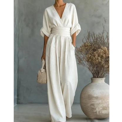 Cream-white jumpsuit on a model, cropped closer to show the V-neckline, pleated waistband and pocket, with dried flowers in a vase. 6