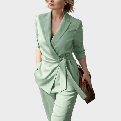 Close three-quarter view of the green wrap blazer knotted at the waist, worn with trousers by a smiling model carrying a brown bag. 6