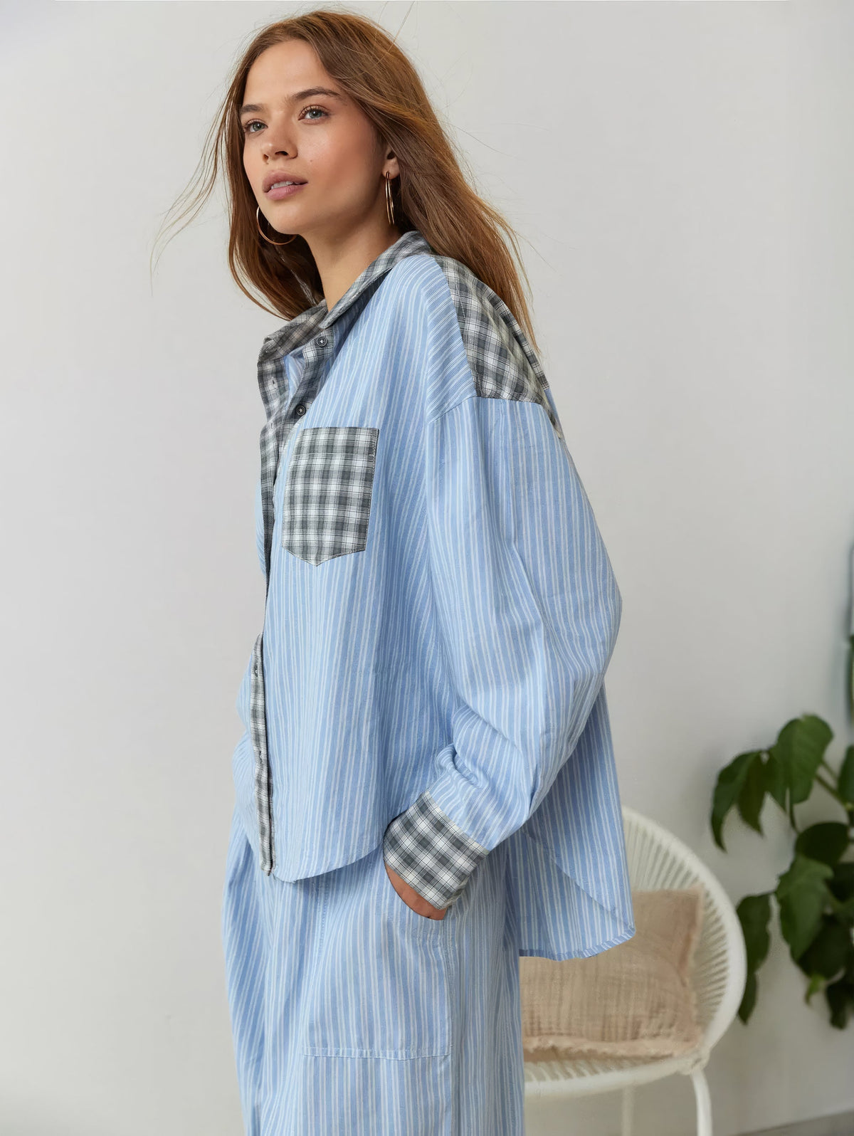 Side view of the blue striped pyjama set on a model, with grey check pocket, cuffs and shoulder panels, plant and chair behind. 3