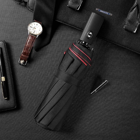 Folded black umbrella with red trim lying on a dark surface beside a wristwatch and pen 1