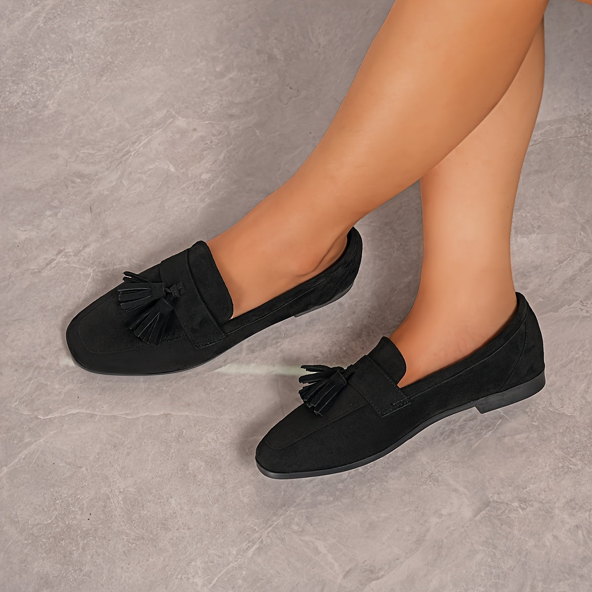 Black tasselled loafers worn with legs crossed, shot from above on a grey stone floor 20