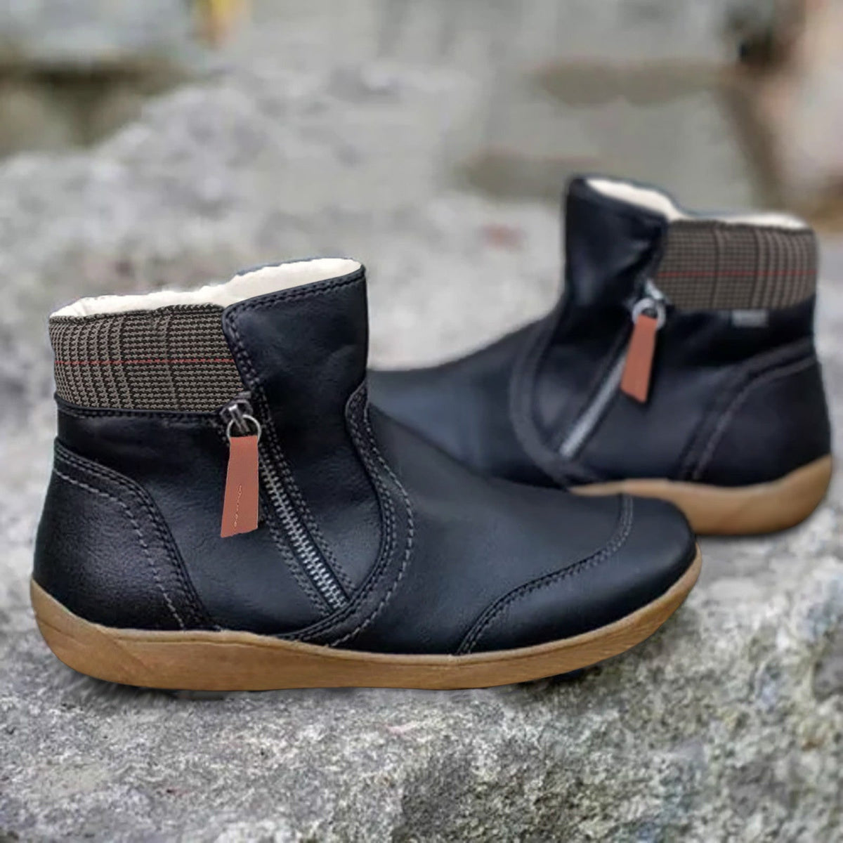 Pair of black leather ankle boots with side zip, orange-brown pull tab, checked collar and fleecy lining, on a stone surface. 2