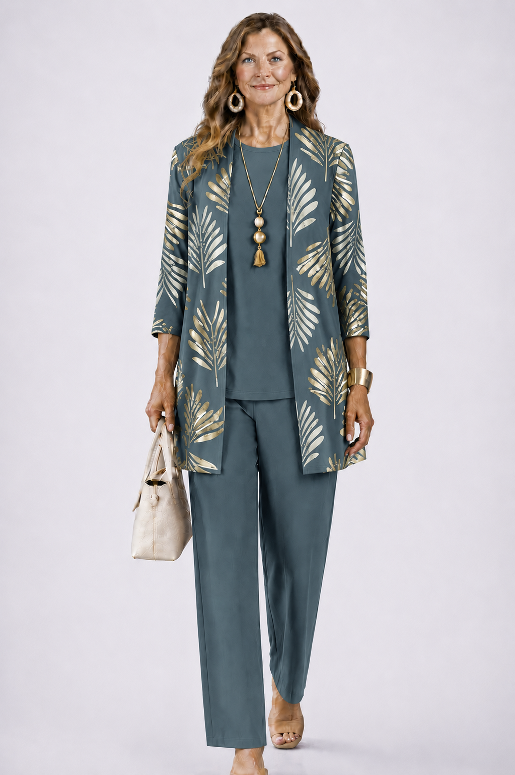 Woman wearing the set in slate blue-grey with gold palm-leaf print on the open cardigan, plain top and trousers, holding a cream handbag. 1