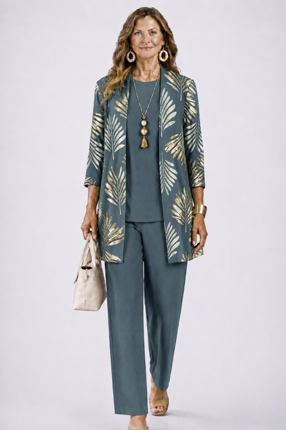 Woman wearing the set in slate blue-grey with gold palm-leaf print on the open cardigan, plain top and trousers, holding a cream handbag. 1