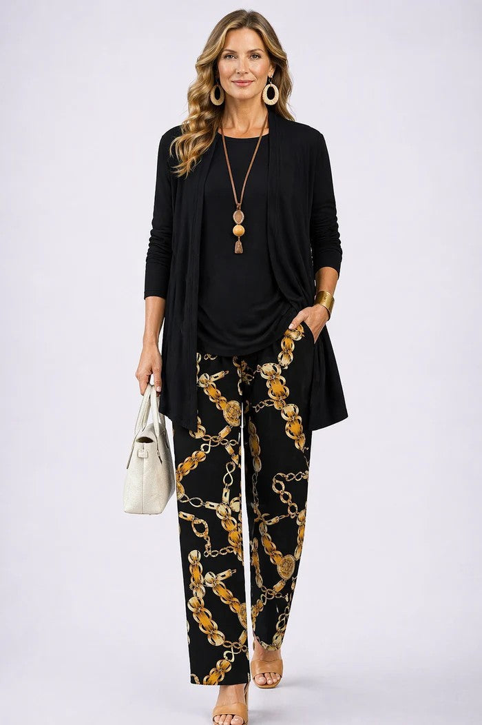Model in a plain black open cardigan and top with black trousers printed with gold chain links, holding a cream handbag. 17