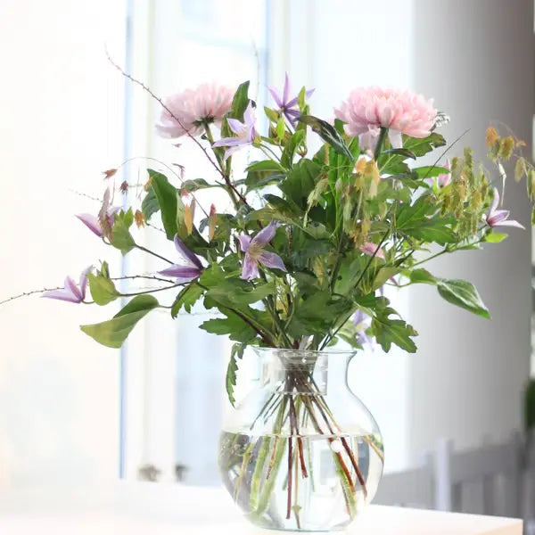 Loose pink and purple flowers with leafy stems arranged in a round clear glass vase of water, beside a bright window. 4