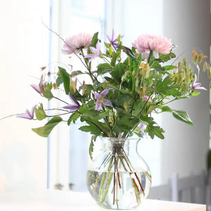 Loose pink and purple flowers with leafy stems arranged in a round clear glass vase of water, beside a bright window. 4