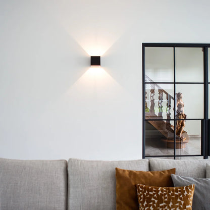 Black cube wall lamp on a plain white wall above a grey sofa with orange cushions, next to a glazed black-framed partition. 10