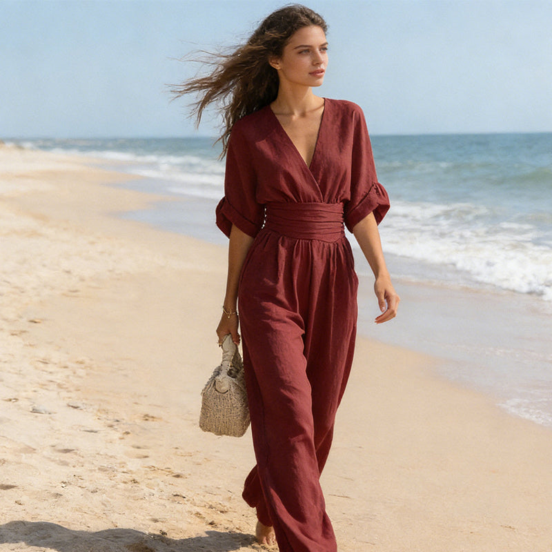 Model in the burgundy jumpsuit walking barefoot along a sandy beach with the sea behind, hair blowing, carrying a woven bag. 7