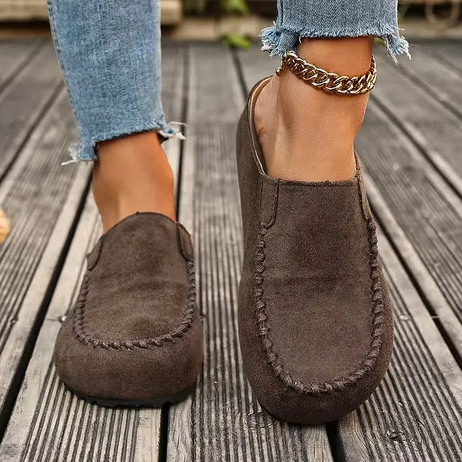 Front view of dark brown slip-on mules worn with frayed jeans on wooden decking, showing the rounded toe and stitched trim. 4