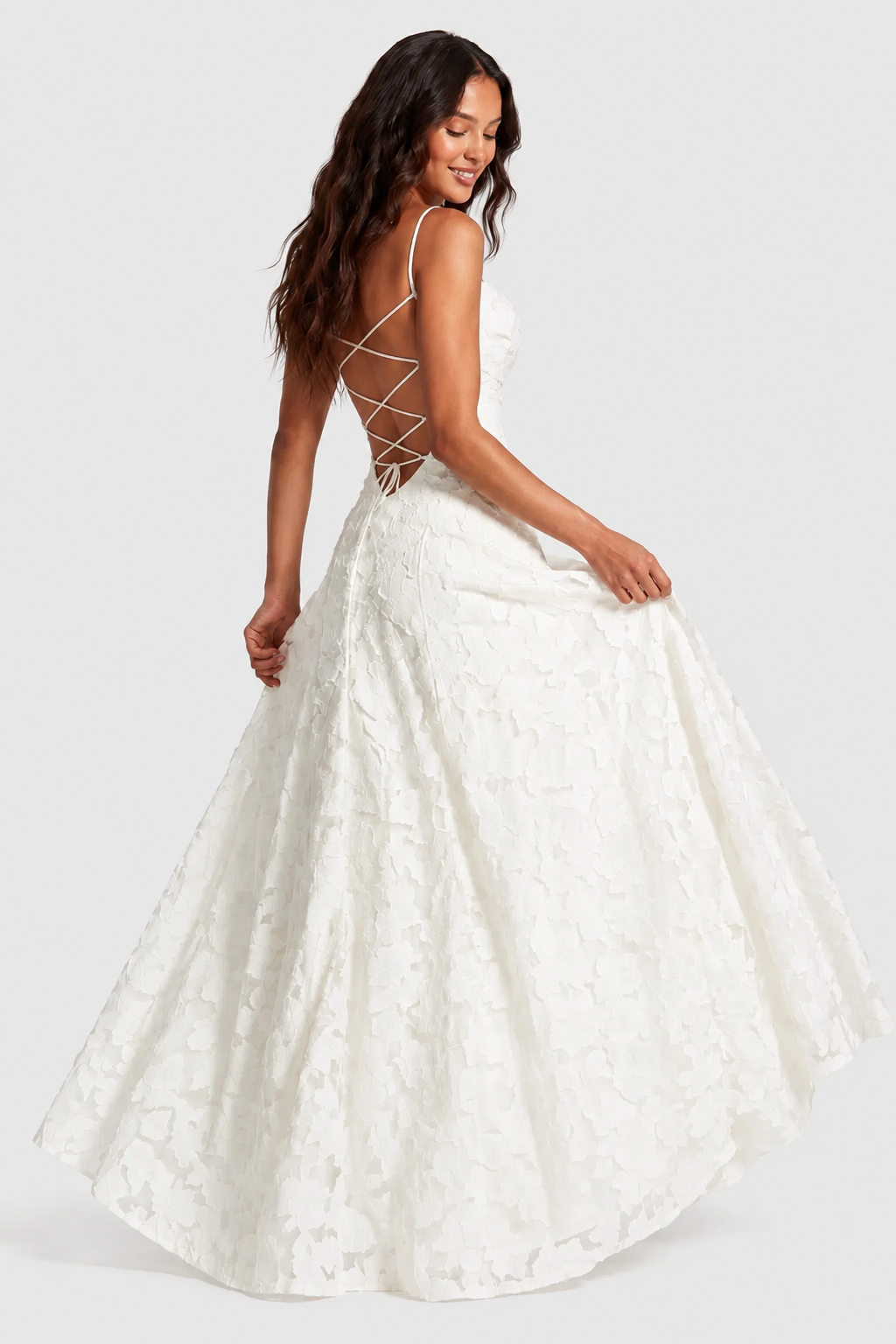 Back view of a white lace gown with criss-cross lace-up back, model holding the skirt out as she glances down. 8