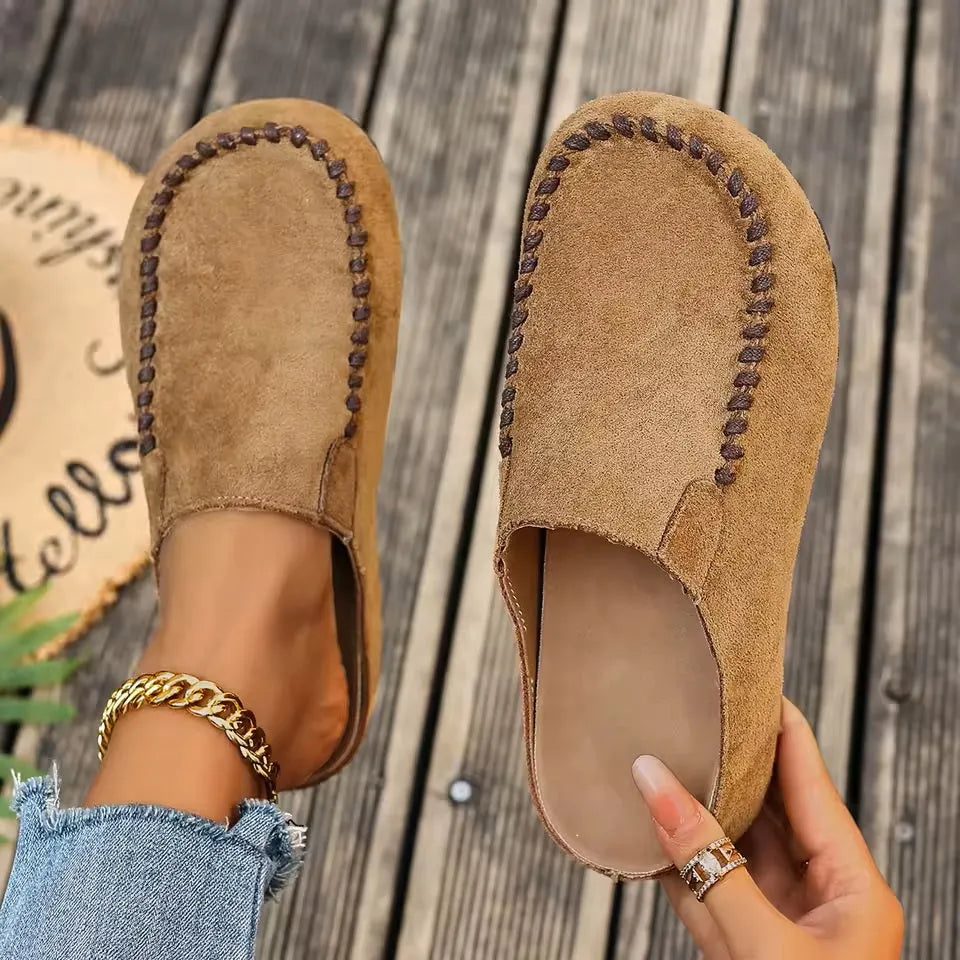 Pair of tan suede-look slip-on mules with contrast stitching, one worn on a foot and one held to show the cushioned insole. 1