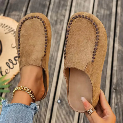 Pair of tan suede-look slip-on mules with contrast stitching, one worn on a foot and one held to show the cushioned insole. 1