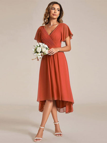 Model in the burnt orange chiffon dress holding a small bouquet of white flowers, standing in white heeled sandals. 95
