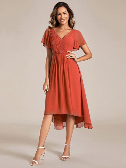 Model in a burnt orange chiffon dress with pleated wrap V-neck, flutter sleeves and high-low hem, one hand on her hip. 92