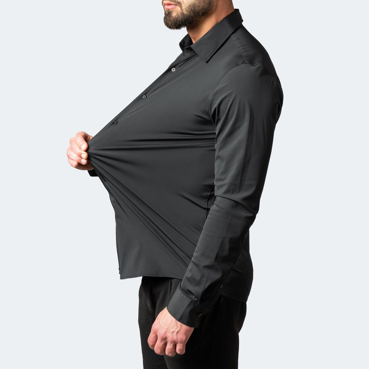 Profile of a bearded model in a black shirt pulling the front outwards to show how far the fabric stretches. 2