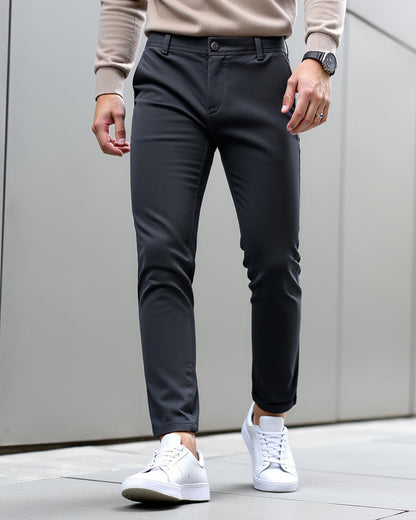 Charcoal chinos worn with a beige knit jumper, black watch and white trainers against a grey panelled wall. 9