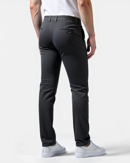 Back view of charcoal chinos on a model, showing a buttoned back pocket, belt loops and white trainers. 8