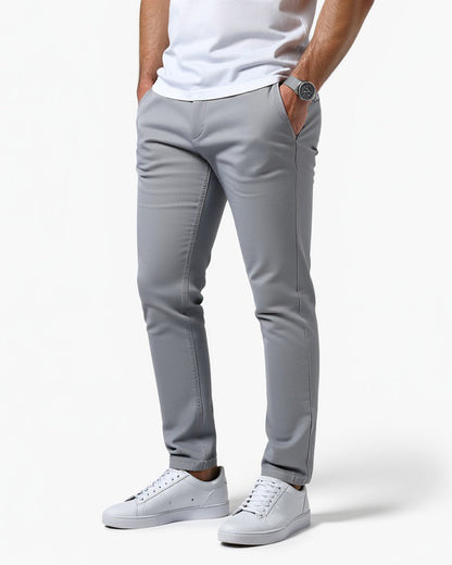 Light grey slim chinos worn by a model with a white t-shirt, white trainers and a watch, one hand in a pocket. 1