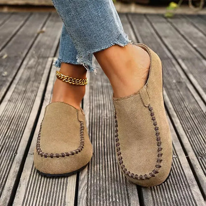 Front-angled view of the sandy beige slip-ons worn on wooden decking, showing the closed toe, brown stitching and open heel. 5