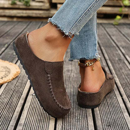 Side and back view of the brown backless mules worn on decking, showing the flat black sole and open heel. 6