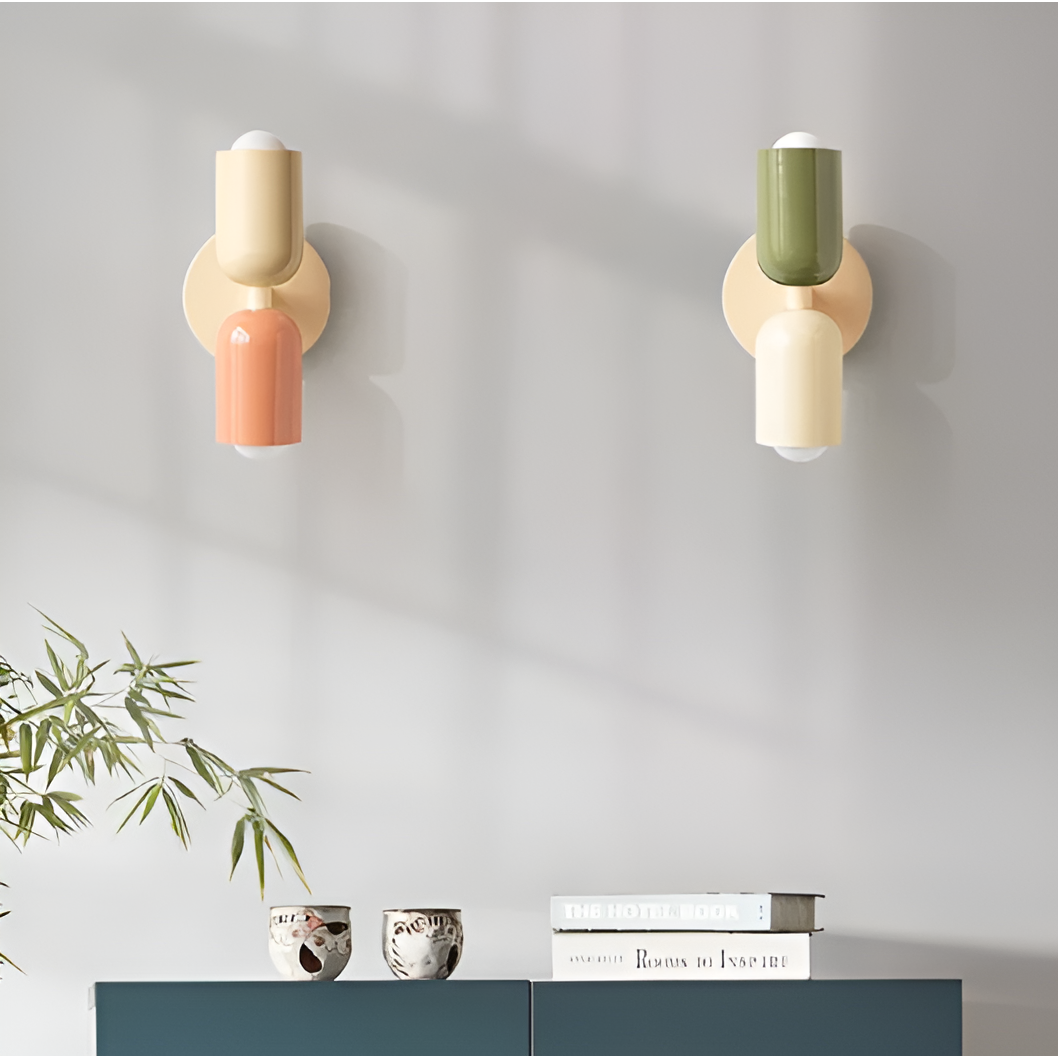 Two twin-head wall lights mounted on a pale wall above a blue-green sideboard, one cream and peach, the other green and cream. 2
