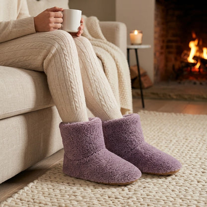 Mauve plush ankle slippers worn with cream cable-knit leggings on a sofa, with a mug, blanket and lit fireplace nearby. 2