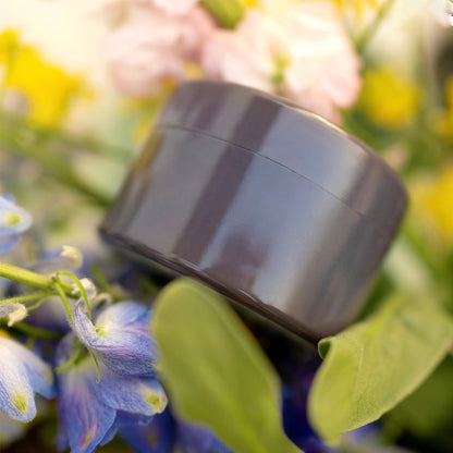 Glossy grey round insert with a lid seam, surrounded by blue flowers, green leaves and blurred pink and yellow blooms. 16