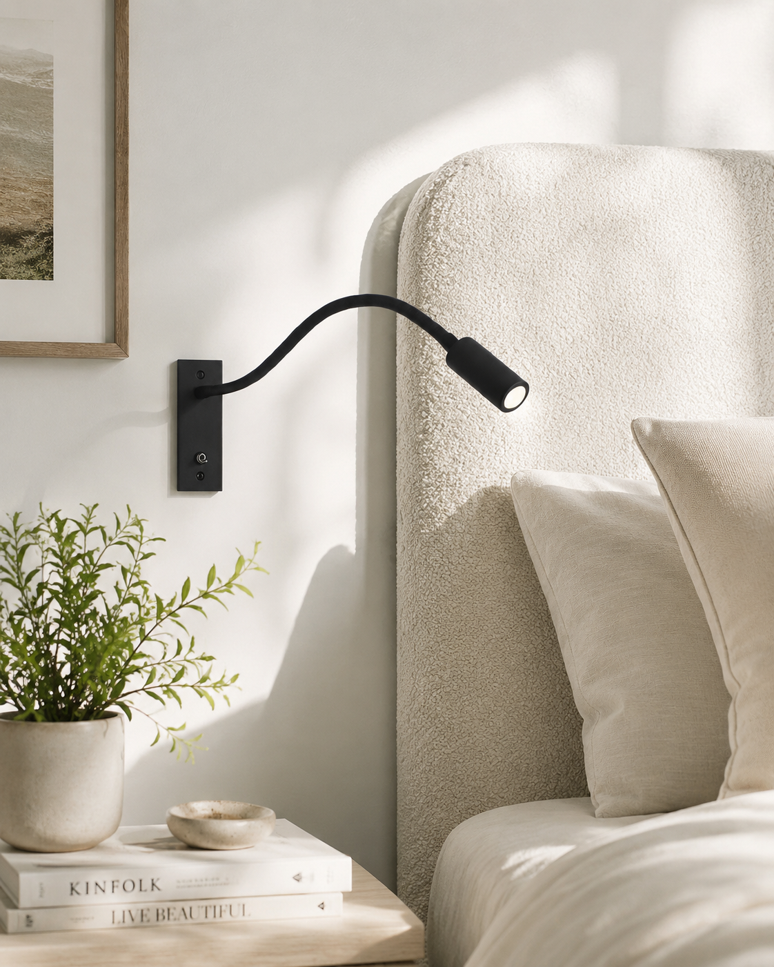 Black flexible wall light mounted beside a cream bouclé headboard, with a plant and books on the bedside table. 2