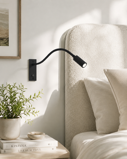 Black flexible wall light mounted beside a cream bouclé headboard, with a plant and books on the bedside table. 2