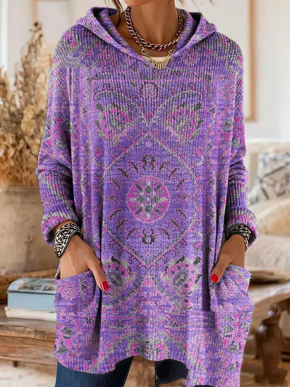 Hooded tunic in purple with a pink and green medallion print, worn by a model with hands in the front pockets. 2