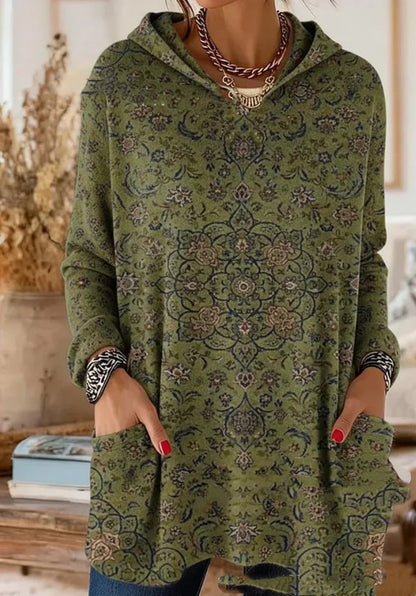 Hooded tunic in olive green with a fine dark floral medallion print, worn by a model with hands in the front pockets. 7