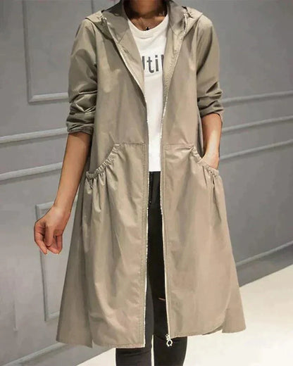 Model wearing the hooded raincoat in light khaki, open over a white top, one hand in a gathered pocket 1