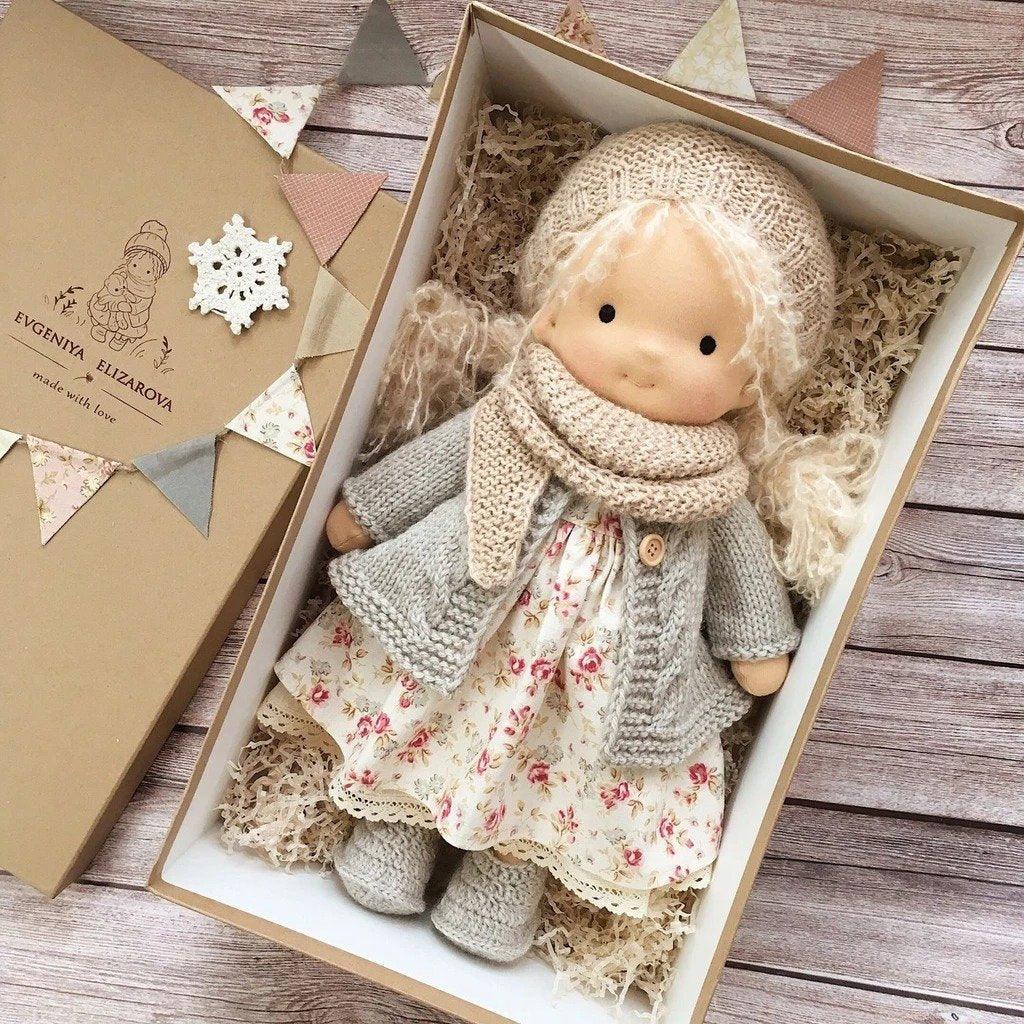 Doll in beige knitted hat and scarf, grey cardigan and floral dress, boxed beside lid with bunting 6