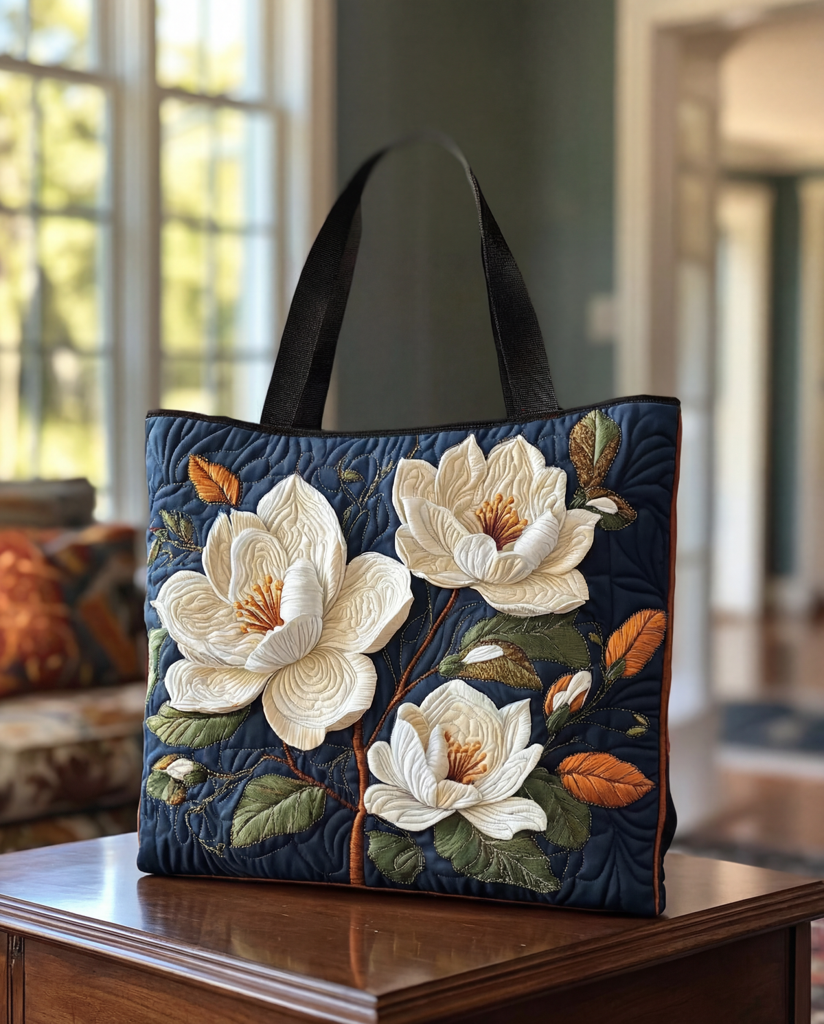 Navy quilted tote with black handles, decorated with raised cream magnolia flowers and green and orange leaves, standing on a polished wooden table. 1