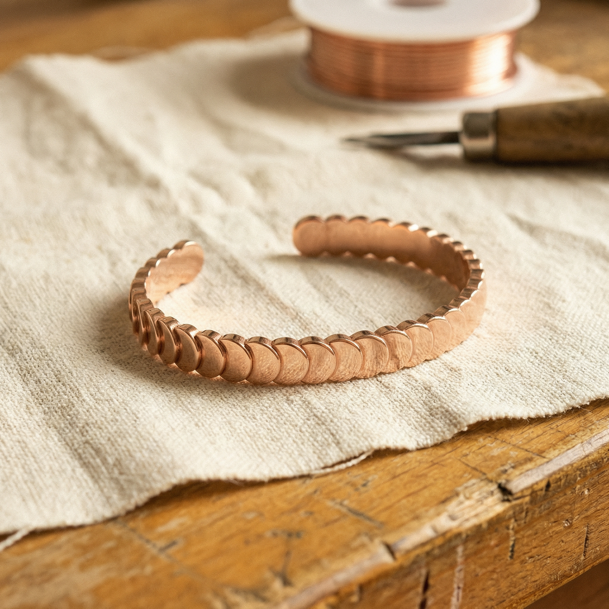 Open copper cuff of overlapping scale discs resting on linen, with a copper wire spool behind 1
