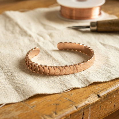 Open copper cuff of overlapping scale discs resting on linen, with a copper wire spool behind 1