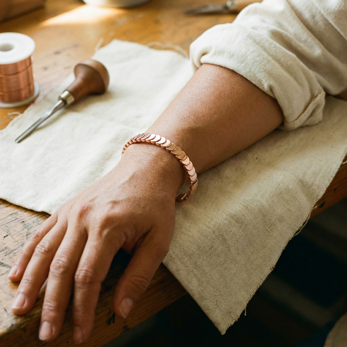 Wrist wearing the open copper scale cuff, hand resting on linen beside a spool and carving tool 2