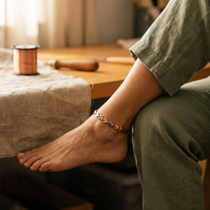 Copper anklet with leaf-shaped links worn on a bare ankle, the wearer seated in green linen trousers beside a spool of copper wire. 1