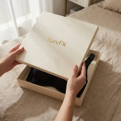 Hands lifting the lid of a cream gift box to reveal the black bra folded inside, resting on a linen bed. 14