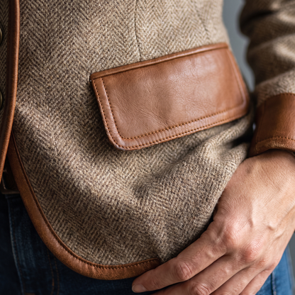Detail of a tan-trimmed pocket flap with contrast stitching on the herringbone tweed, with the model's hand resting nearby. 5