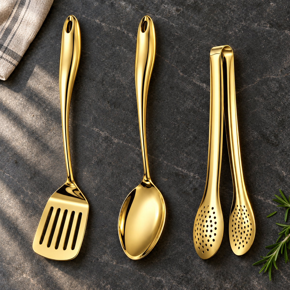 Gold-coloured slotted turner, solid spoon and perforated tongs laid on a dark stone worktop with a striped cloth and rosemary. 23