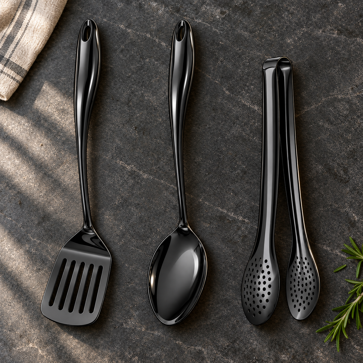 Black slotted turner, solid spoon and perforated tongs laid on a dark stone worktop with a striped cloth and rosemary sprig. 27
