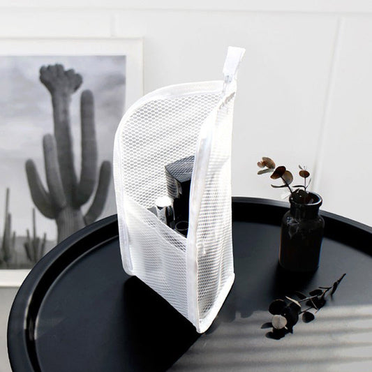 White mesh pouch standing upright on a round black side table, with toiletries visible through the open front pocket and a cactus print behind. 2